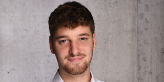 Das Bild ist ein professionelles Porträt eines jungen Mannes vor einer hellgrauen, strukturierten Betonwand. Er hat einen hellen Hautton, dunkelbraune Augen und dunkelbraunes Haar, das an den Seiten kurz geschnitten ist und am Oberkopf länger sowie leicht gewellt fällt. Er trägt einen gepflegten, kurz getrimmten Bart, der sich um den Mund und das Kinn zieht, sowie leichte Stoppeln an den Wangen. Der Mann ist mit einem weißen Langarmhemd bekleidet, dessen oberster Knopf geöffnet ist, was einen legeren, aber ordentlichen Eindruck vermittelt. Sein Oberkörper ist leicht nach rechts gedreht, während sein Kopf frontal zur Kamera ausgerichtet ist. Er blickt den Betrachter direkt an und zeigt ein geschlossenes, freundliches und entspanntes Lächeln.