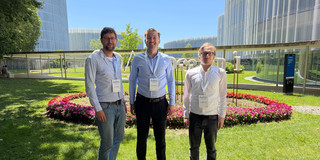The image shows three men standing side-by-side outdoors on a green lawn on a sunny day, looking directly at the camera. All three are wearing long-sleeved button-down shirts, long trousers, and lanyards with name badges clearly displaying the text "ECIS". The man on the left wears glasses, has a slight beard, and is dressed in a light blue shirt, blue jeans, and brown shoes. The man in the middle is smiling and wears a light blue shirt, dark trousers, a brown belt, and brown shoes. The man on the right wears glasses, a white shirt, dark trousers, and black shoes. Behind the men is a circular flower bed with an outer border densely planted with bright pinkish-red flowers. Directly behind the flower bed, large white sculptural letters stand on the grass, though they are partially obscured by the people. The background is dominated by modern buildings featuring striking, undulating white slatted facades that contrast against a clear, bright blue sky. A covered pedestrian walkway is visible in the background on the left, while a dark blue directional signpost stands on the right. In the bottom left corner of the foreground, a small sign is visible showing a red circle with a slash through a dog, indicating that dogs are not permitted.