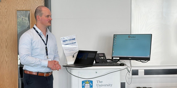 A man with short-cropped light hair and a mustache stands in a bright room, likely a seminar room or office at the University of Sheffield. He is wearing a light blue, finely striped button-down shirt tucked into dark trousers with a brown leather belt. Around his neck, he wears a blue lanyard printed with "The University of Sheffield" holding an ID badge. The man has his hands loosely clasped in front of him and is looking attentively to the right, as if watching a presentation or listening to someone. In front of him is a white mobile lectern featuring the blue and gold crest of the University of Sheffield on its front panel. On top of the lectern are an open black laptop, a black computer keyboard, a mouse, a black desk phone, and a Dell monitor. The monitor displays a university login screen with a reference to "Windows 11." In the background, to the left, there is a light wood door with a narrow vertical glass pane revealing a glimpse of trees outside. A notice with technical instructions is taped to the white wall behind the laptop.