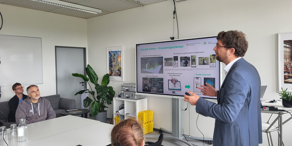 Das Foto im Querformat zeigt eine Präsentationssituation in einem hellen, modernen Seminar- oder Besprechungsraum. Auf der rechten Bildseite steht ein männlicher Referent im Profil, der den Blick nach links zu den Zuhörenden gerichtet hat. Er hat kurzes, lockiges braunes Haar, trägt eine Brille, ein blaues Sakko über einem weißen Hemd sowie eine blaue Jeans. Er gestikuliert mit beiden Händen, wobei er in der rechten Hand einen Presenter hält. Hinter ihm steht ein großer, digitaler Flachbildschirm auf einem mobilen Rollständer. Die darauf angezeigte Präsentationsfolie trägt die grüne Überschrift „Virtuelle Gärten – Einsatzmöglichkeiten“ und zeigt oben rechts das Logo der TU Dortmund. Der Inhalt der Folie ist in vier Bereiche unterteilt, die Bilder und Grafiken zu Augmented Reality (AR) und Virtual Reality (VR) Anwendungen auf Smartphones und Computerbildschirmen darstellen.  Auf der linken Bildhälfte sitzen die Zuhörenden an einer Anordnung aus weißen Tischen. Im direkten Vordergrund, mit dem Rücken zur Kamera, ist der Kopf einer Person mit rötlich-braunem, zum Dutt gebundenen Haar zu sehen, die einen schwarzen Pullover trägt. Dahinter sitzt ein Mann mit grau meliertem Haar und Bart in einem grauen Kapuzenpullover, der aufmerksam zum Referenten blickt. Vor ihm auf dem Tisch stehen eine gläserne Wasserkaraffe und mehrere Trinkgläser. Links im Hintergrund sitzt ein weiterer Mann mit Brille und blauem Hemd auf einem dunkelgrauen Sofa. Der Raum ist zweckmäßig eingerichtet: Im Hintergrund erkennt man ein weißes Regal, in dem silberne Koffer und gelbe Stapelboxen lagern, sowie eine große grüne Zimmerpflanze (Monstera) in der Ecke. An den weißen Wänden hängen gerahmte Bilder, und an der Decke sind längliche Leuchtstoffröhren angebracht.  English The landscape-format photo captures a presentation taking place in a bright, modern seminar or meeting room. On the right side of the image, a male presenter stands in profile, facing left towards the audience. He has short, curly brown hair, wears glasses, a blue blazer over a white shirt, and blue jeans. He is gesturing with both hands whilst holding a presentation clicker in his right hand. Behind him is a large digital flat-screen display mounted on a rolling stand. The slide being presented features the green headline "Virtuelle Gärten – Einsatzmöglichkeiten" (Virtual Gardens – Application Possibilities) and shows the logo of TU Dortmund in the top right corner. The slide content is divided into four quadrants displaying images and graphics related to Augmented Reality (AR) and Virtual Reality (VR) applications on smartphones and computer monitors.  On the left side of the image, the audience is seated at a configuration of white tables. In the immediate foreground, with their back to the camera, is the head of a person with reddish-brown hair tied in a bun, wearing a black sweater. Further back sits a man with grey-flecked hair and a beard, wearing a grey hoodie, who is looking attentively at the speaker. A glass water carafe and several glasses are placed on the table in front of him. In the background on the far left, another man wearing glasses and a blue shirt is seated on a dark grey sofa. The room is functionally furnished: in the background, a white shelving unit holding silver cases and yellow stacking bins is visible, alongside a large green houseplant (Monstera) in the corner. Framed pictures hang on the white walls, and long fluorescent light fixtures are mounted on the ceiling.