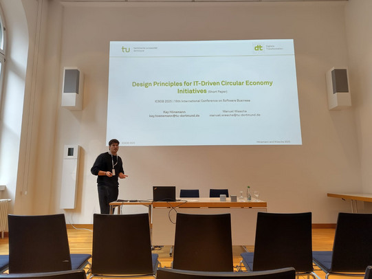 Das Foto zeigt eine Präsentationssituation in einem hellen Konferenzraum mit hoher Decke und Holzboden. Im Zentrum des Bildes hängt eine große weiße Projektionsleinwand, auf der die Titelfolie eines Vortrags zu sehen ist. Oben links auf der Folie befindet sich das grüne Logo der "tu technische universität dortmund", oben rechts das Logo "dt Digitale Transformation". Der Titel der Präsentation lautet in grüner Schrift "Design Principles for IT-Driven Circular Economy Initiatives (Short Paper)". Darunter steht in kleinerer grauer Schrift "ICSOB 2025 | 16th International Conference on Software Business". Im unteren Teil der Folie sind die Autoren Kay Hönemann und Manuel Wiesche mit ihren E-Mail-Adressen der TU Dortmund aufgeführt.  Links neben der Leinwand steht ein männlicher Referent mit dunklen Haaren und Brille. Er trägt einen schwarzen Pullover, eine dunkle Hose und ein weißes Umhängeband (Lanyard) um den Hals. Er hält einen Presenter in der Hand, gestikuliert leicht und blickt zum Publikum. Vor der Leinwand steht ein heller Holztisch, auf dem ein aufgeklappter Laptop, diverse Kabel, zwei kleine Lautsprecher sowie Wasserflaschen und Gläser stehen. An der linken Wand befinden sich hohe Fenster und weiße Lautsprecherboxen.