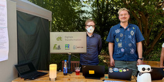 Zwei Männer stehen im Freien hinter einem Informationsstand aus Holz. Der linke Mann trägt eine FFP2-Maske, der rechte Mann ein dunkelblaues Hemd mit hellen Mustern. In der Mitte des Tisches zeigt ein Monitor das Logo „dt Digitale Transformation“ mit drei kleineren Projektlogos. Auf dem Tisch stehen zusätzlich zwei Laptops, eine orangefarbene Lampe, Informationsflyer und ein kleiner weißer humanoider Roboter.