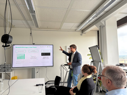 Das Foto zeigt eine Szene während eines Workshops oder einer Präsentation in einem hell beleuchteten Seminarraum. Im Mittelpunkt steht ein Mann, der Referent, der rechts neben einem großen, auf einem Ständer montierten Flachbildschirm steht. Er trägt eine Brille, ein blaues Sakko über einem hellen Hemd sowie Jeans und gestikuliert mit seiner rechten Hand in Richtung der Präsentation, während er Blickkontakt zu den Zuhörenden hält. Auf dem Bildschirm ist eine Folie mit der grünen Überschrift „Herausforderungen in der Praxis“ zu sehen. Die Folie zeigt Logos der TU Dortmund und „dt“, ein grünes Balkendiagramm auf der linken Seite, ein Zitat in der Mitte sowie rechts ein technisches Bild (vermutlich Augmented-Reality-Hardware) mit Spezifikationen wie Sichtfeld und Gewicht.  Im rechten Vordergrund sitzen zwei Personen im Profil, die dem Vortrag aufmerksam folgen: Ganz rechts ein Mann mit kurzem grauen Haar, Brille und hellblauem Hemd, daneben eine Frau mit rötlich-braunem, hochgestecktem Haar, schwarzem Oberteil und einem gelben Kragen oder Tuch. Der Raum wirkt funktional eingerichtet; an der Rasterdecke befinden sich längliche Leuchtstoffröhren. Links im Bild hängt ein schwarzer Würfel mit Steckdosen an einer Kette von der Decke herab, darunter steht ein weißes Regal mit mehreren silbernen Metallkoffern. Rechts hinter dem Referenten stehen ein Flipchart und eine Zimmerpflanze vor einer großen Fensterfront, die den Blick auf einen bewölkten Himmel und eine Stadtlandschaft freigibt. Auf dem weißen Tisch im direkten Vordergrund stehen Wassergläser und ein Smartphone liegt bereit.