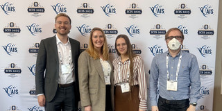 The image is a full-body group photo showing four people standing side-by-side facing the camera in front of a conference backdrop. The backdrop is white and features a banner at the top reading "ICIS 2025" in large dark blue letters, followed by "Nashville Tennessee December 14 – 17". The banner includes graphics of a guitar headstock on the left and an orange guitar neck on the right containing the hashtag #ICIS2025. Below the banner, the wall features a repeating step-and-repeat pattern of logos for "AIS" (Association for Information Systems) and "ICIS 2025".  From left to right: The first person is a man with short blond hair and glasses, wearing a dark grey suit jacket, a white button-down shirt, dark trousers, and brown leather dress shoes. Next to him is a woman with long blond hair wearing a beige checked blazer, a white top, black trousers, and light-colored sneakers. Beside her stands a woman with long brown hair and glasses, wearing a vertically striped shirt in white and reddish-brown, a brown midi skirt, dark tights, and tan boots with fur trim. On the far right is a man with short hair and glasses wearing a white face mask. He is dressed in a blue checkered button-down shirt, dark jeans, and brown boots. All four individuals are wearing conference name badges on lanyards around their necks. They are standing on a carpet patterned with large, abstract leaf shapes in shades of blue and green.