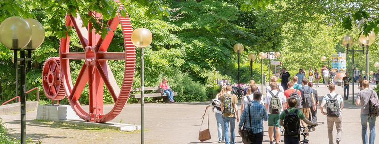 Students walk along a tree-lined path on the North Campus in summer, passing a sculpture with large red gears.