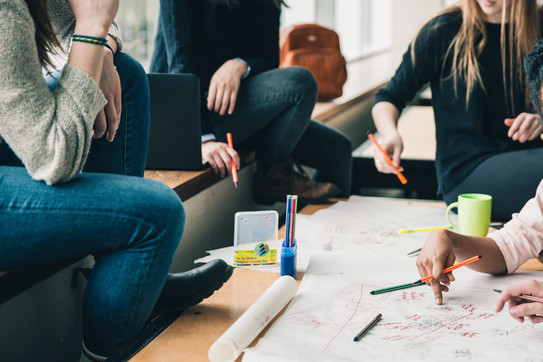Brainstorming Vier Menschen sitzen an einem Tisch, die Gesichter sind nicht im Bild. Sie halten Stifte in der Hand und sehen nachdenkend aus. Eine Hand schreibt etwas auf das Plakat auf dem Tisch, welches sehr unstrukturiert gestaltet ist. Auf dem Tisch sind noch mehr Stifte und eine Tasse. Auf der Sitzbank erkennt man einen geöffneten Laptop und einen Rucksack im Hintergrund.
